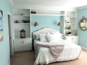 Un dormitorio con cama, mesita de noche, estante con objetos decorativos y una puerta.
