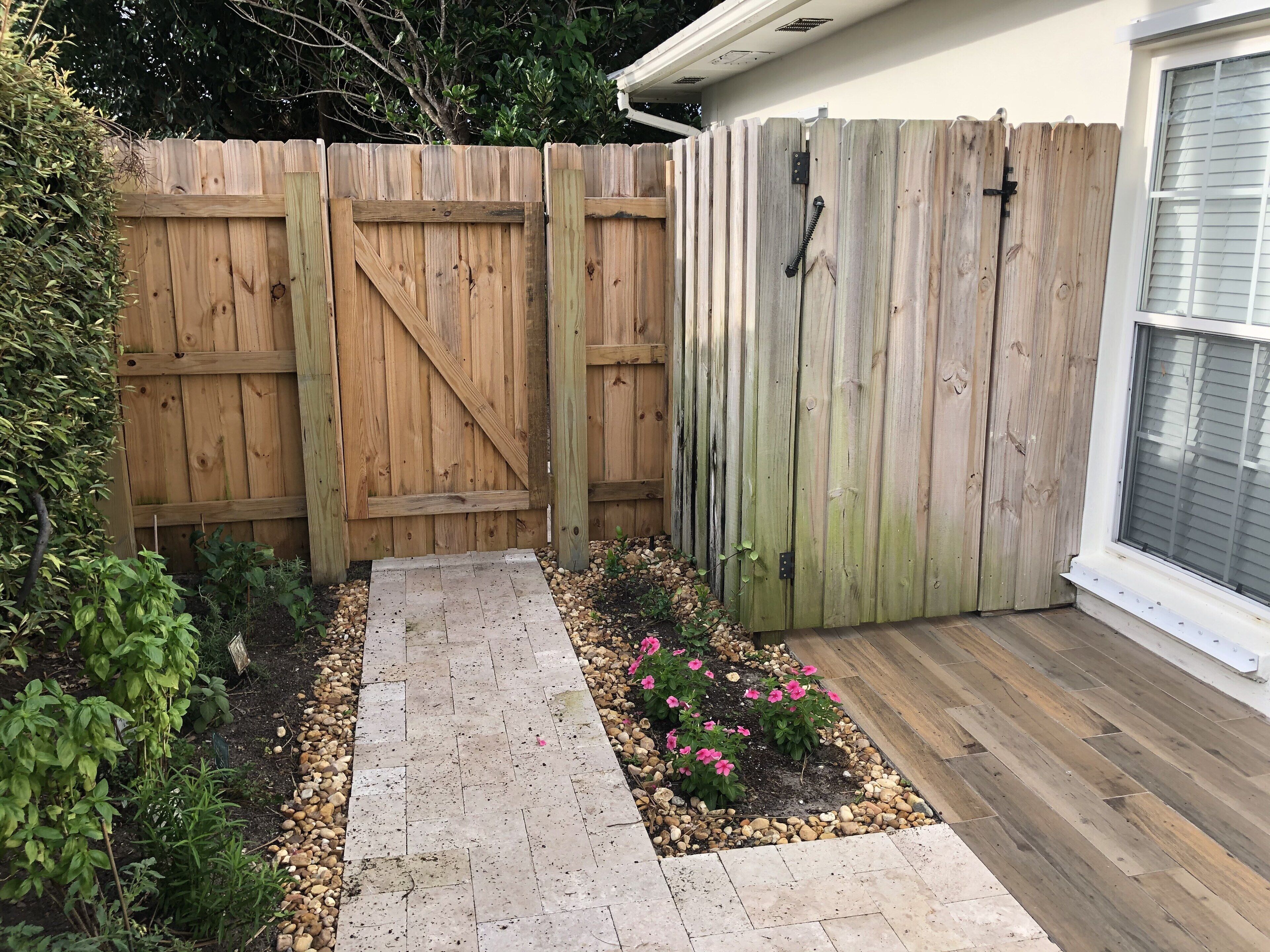 Una valla de madera con un portón, un camino pavimentado y un jardín con flores rosas.