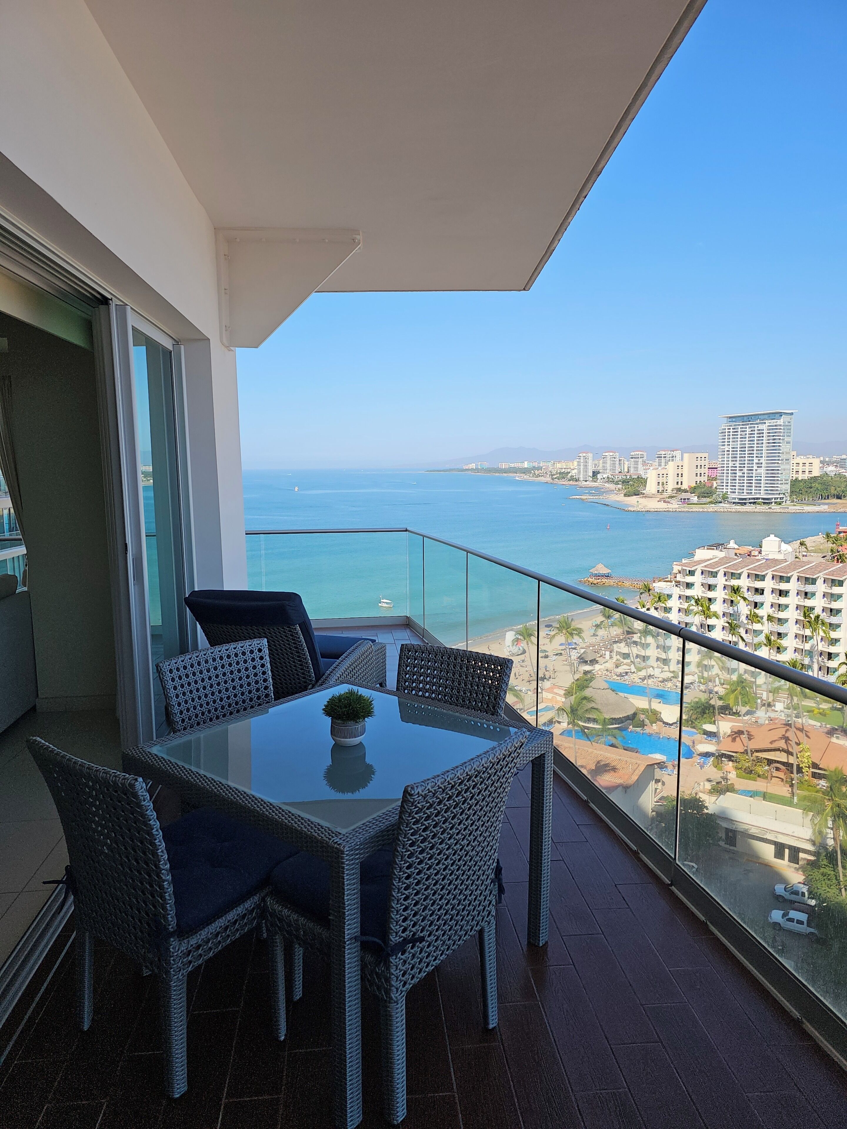 A balcony with a glass table and wicker chairs, overlooking a beach and ocean.