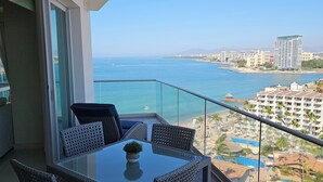 Ein Balkon mit Glastisch und Korbstühlen, der Blick auf einen Strand und den Ozean geht.