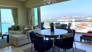 Ein geräumiges Wohnzimmer mit einem großen runden Tisch und blauen Stühlen, ein beiges Sofa mit gemusterten Kissen sowie ein Blick auf ein Kreuzfahrtschiff und eine Stadtlandschaft durch große Fenster.