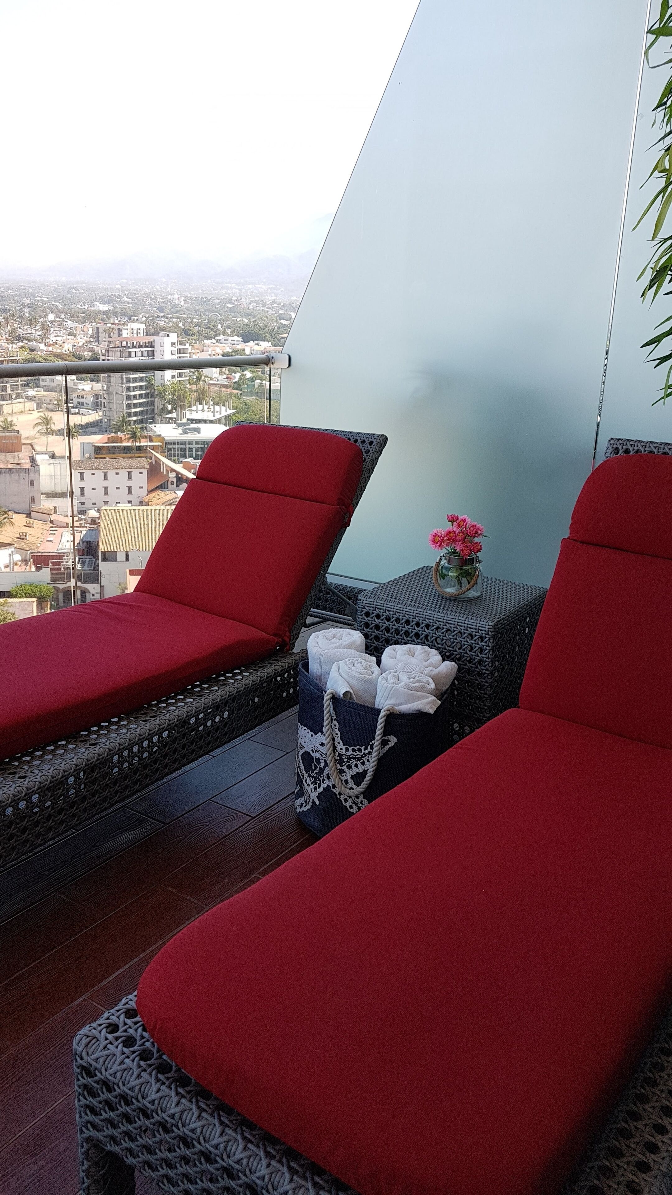 A balcony with two red cushioned chairs, a wicker table with a vase of flowers, and a view of a cityscape.