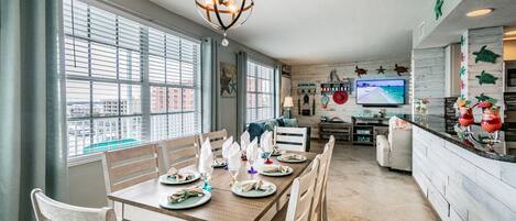 A dining area with a wooden table set for six, a chandelier, and a view of the city through large windows.