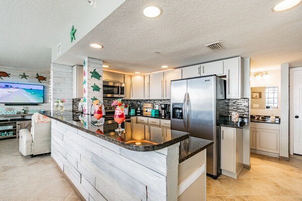 A modern kitchen with white cabinetry, a central island, stainless steel appliances, and a flat-screen TV mounted on the wall.