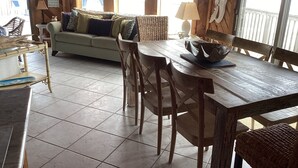 Ein gemütlicher Wohnbereich mit einer Couch, einem Holzesstisch und Stühlen. An der Wand hängt ein Schild und durch große Fenster ist der Blick auf das Meer möglich.