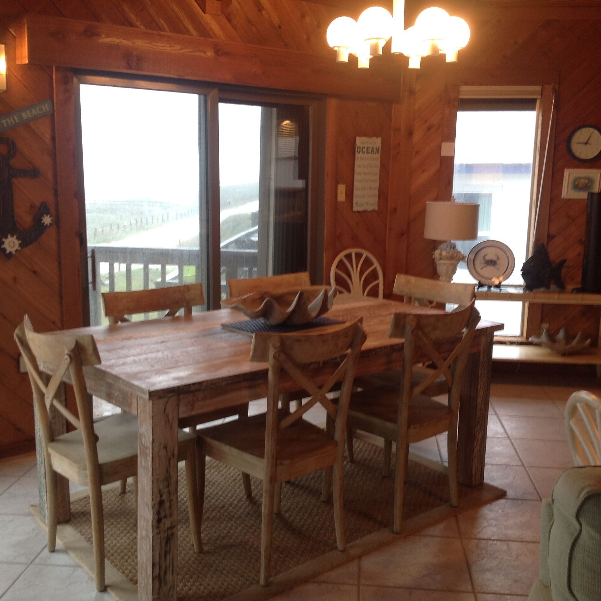 Comedor de madera con una mesa grande y sillas, vista al océano a través de un ventanal, y un reloj en la pared.
