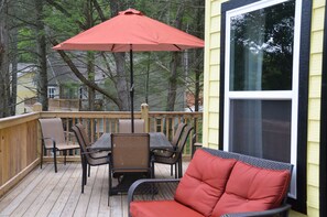 Eine Holzterrasse mit Tisch, Stühlen und einem roten Sonnenschirm sowie einer Korbsitzgruppe.