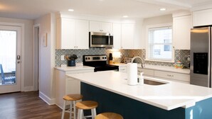 A modern kitchen with white cabinetry, stainless steel appliances, and a central island with stools. - Beach Cottage Multiple Suites & Walk to Boardwalk! (Virginia Beach)