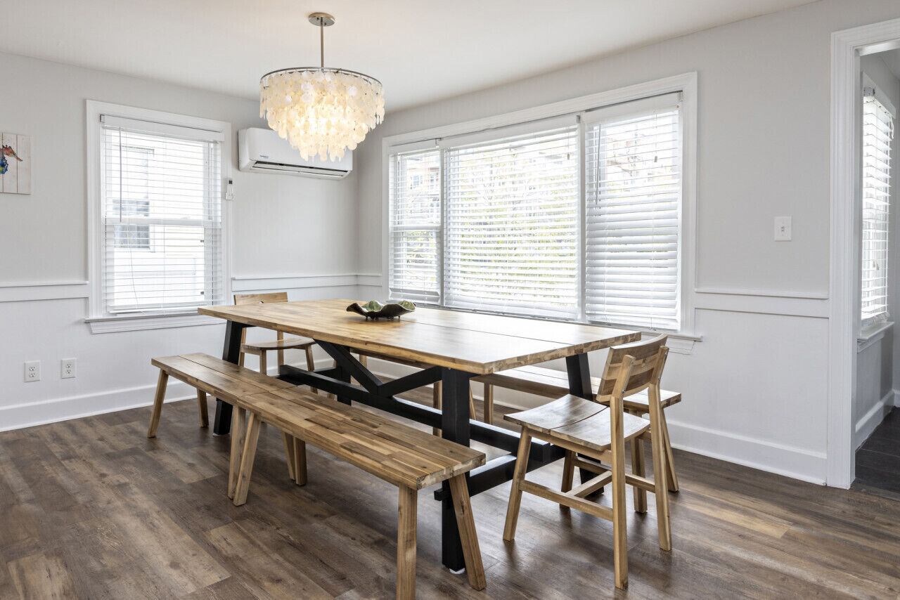 A dining area with a wooden table and benches, large windows with blinds, and a chandelier.