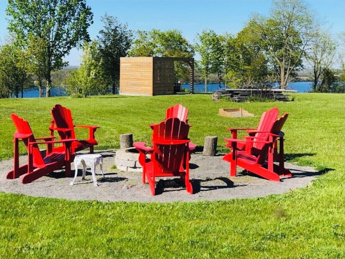 A grassy outdoor area with red Adirondack chairs and a small white table, a wooden structure, and a body of water in the background.