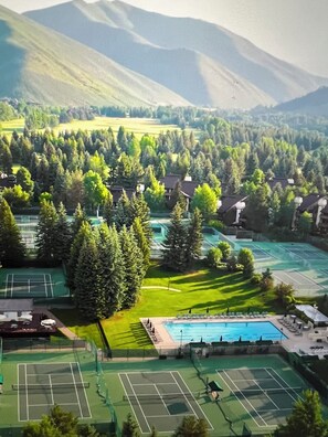A tennis court surrounded by lush greenery and residential buildings, with mountains in the background.
