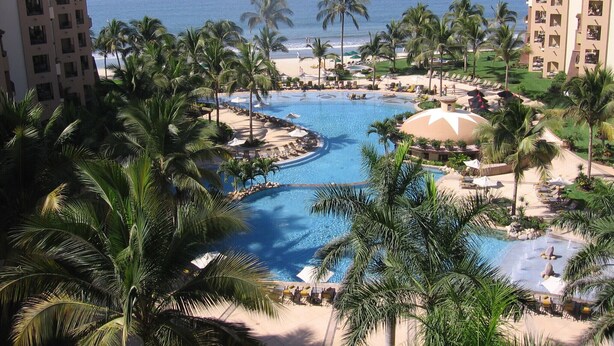 Un resort con piscina central, palmeras y vista al océano.