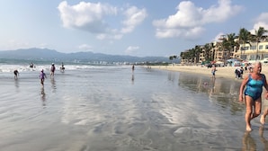 Una playa con gente caminando y nadando, palmeras y edificios al fondo.