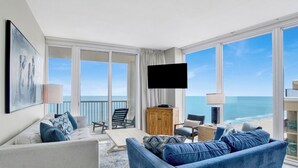 A modern living room with a large window overlooking the ocean, a flat-screen TV, and a cozy seating area with a sofa and armchairs.