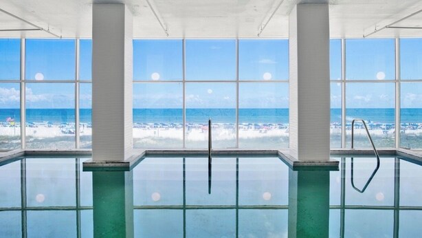 An indoor swimming pool with large windows offering a view of the beach and ocean.
