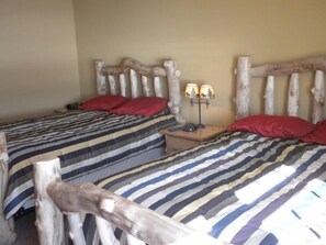Quarto com duas camas, cada uma com colcha listrada e almofadas vermelhas. Há uma mesa de cabeceira com abajur entre as camas.