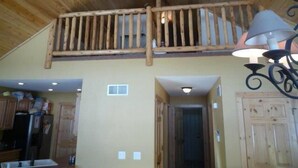 Un intérieur rustique avec un plafond en bois, un balcon doté d’une balustrade, un coin cuisine avec des placards et un couloir menant à une porte.