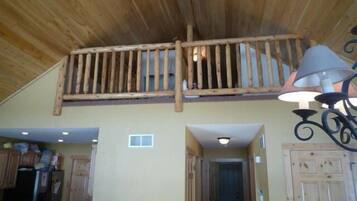 A rustic interior with a wooden ceiling, a balcony with a railing, a kitchen area with cabinets, and a hallway leading to a door.