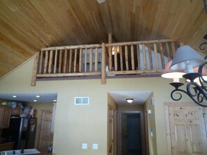 Un intérieur rustique avec un plafond en bois, un balcon doté d’une balustrade, un coin cuisine avec des placards et un couloir menant à une porte.