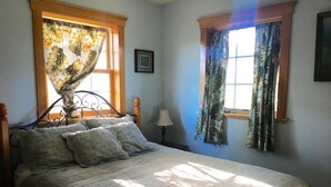 Une chambre à coucher avec un lit, deux fenêtres avec des rideaux, une lampe de chevet et un tableau encadré au mur.