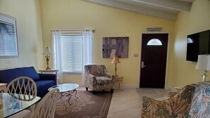A living room with a blue sofa, a patterned armchair, a glass coffee table, and a dining table with chairs. There is a door with a sign above it, a lamp on a side table, and a television mounted on the wall.