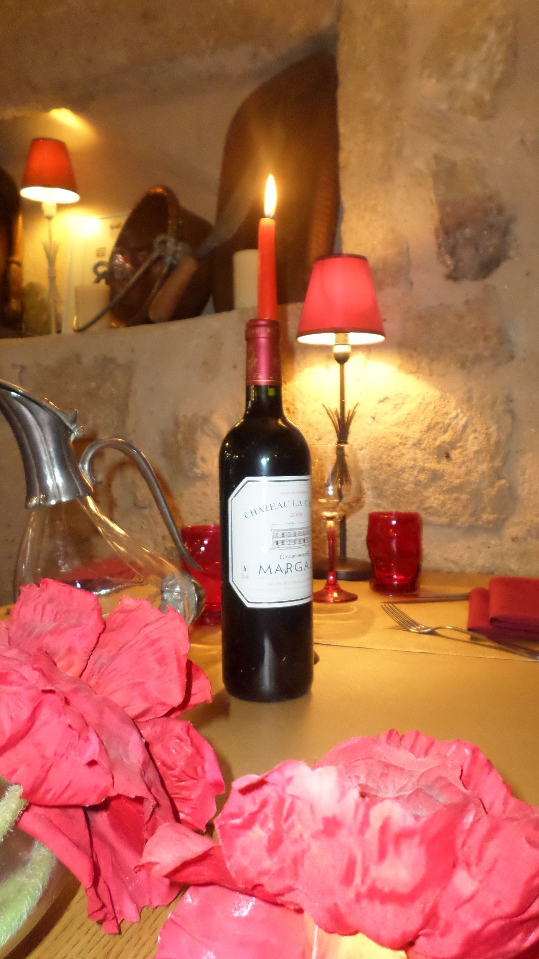 Eine Flasche Chateau La Garde Margaux 2008 Wein, eine Kerze und rote Blumen auf einem Tisch mit einer Wand aus Stein im Hintergrund.