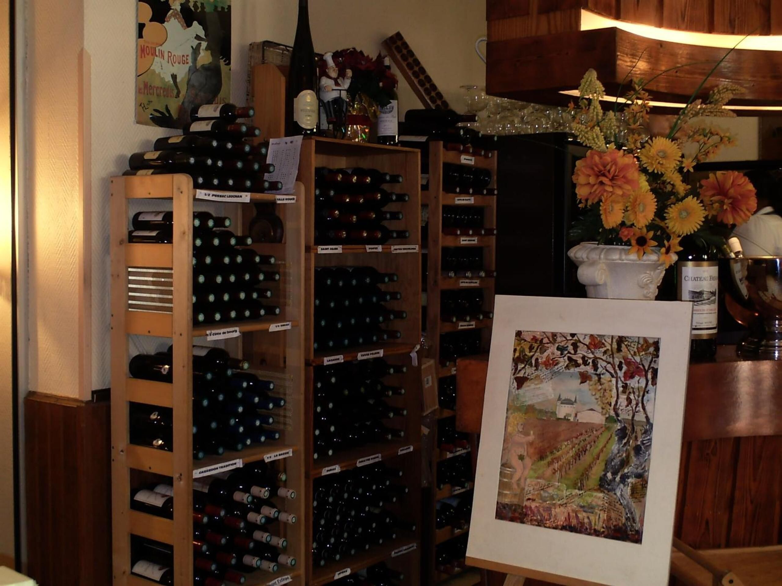 A wine cellar with wooden racks filled with wine bottles, a painting of a vineyard, and a vase of flowers on a counter.