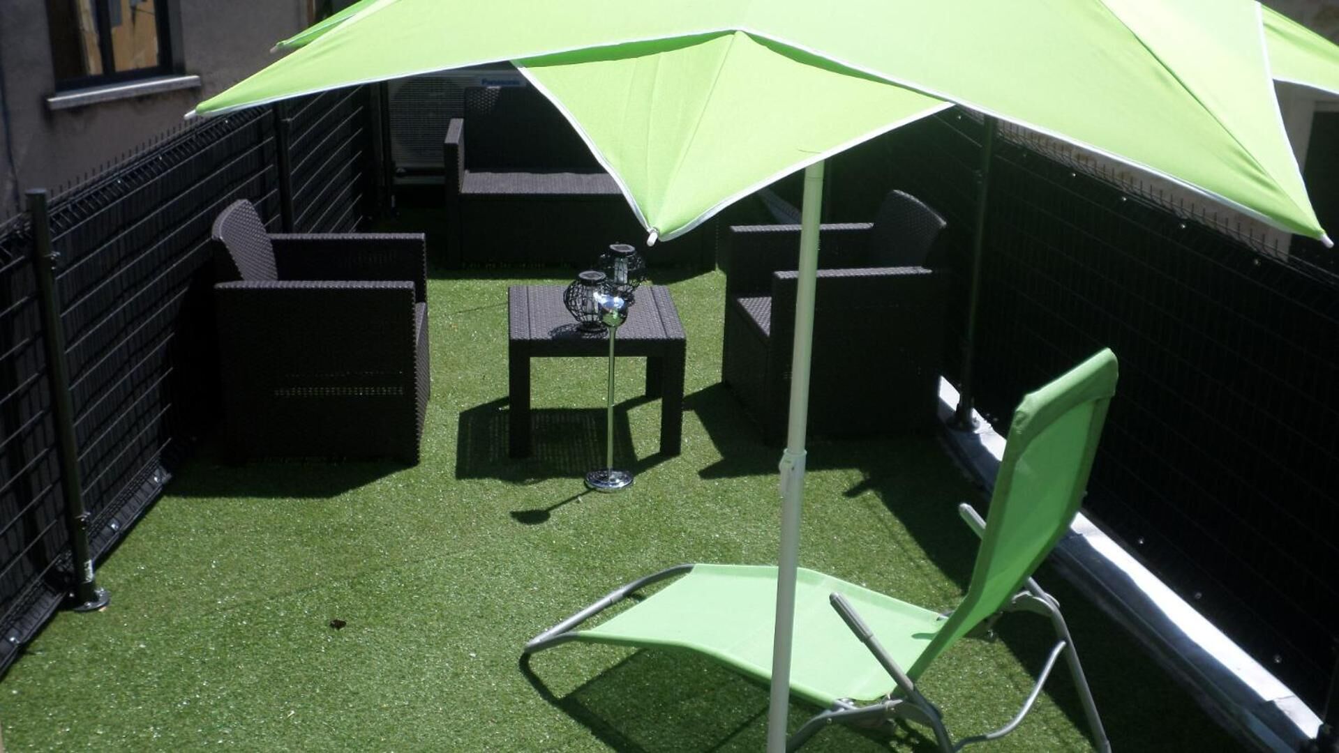 Un espace patio avec une pelouse artificielle, un parasol vert, une table agrémentée d’un objet décoratif et du mobilier d’extérieur.