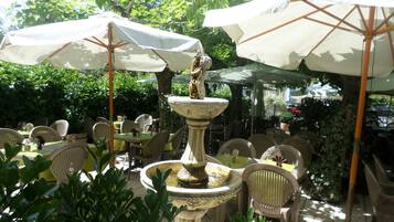 An outdoor dining area with tables and chairs, umbrellas, and a central fountain.
