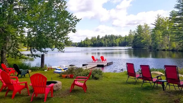A lakeside deck with red chairs and a dock extending into the water. - Balsam Point Lakehouse · Indian Lake Adirondacks · Waterfront on Lake Abanakee (Indian Lake)
