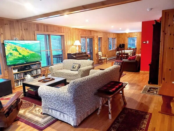 A cozy living room with wooden walls, a large flat-screen TV, a sofa set, a dining table, and a red accent wall.