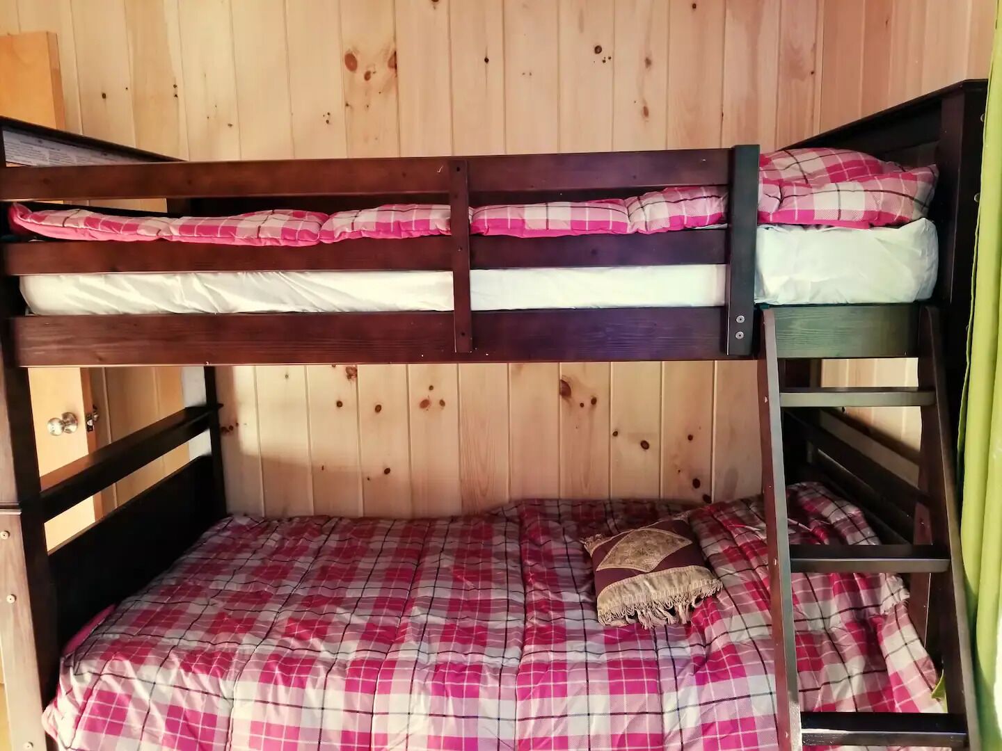 A bunk bed with a checkered bedspread and pillows, wooden walls, and a small shelf with a pillow.