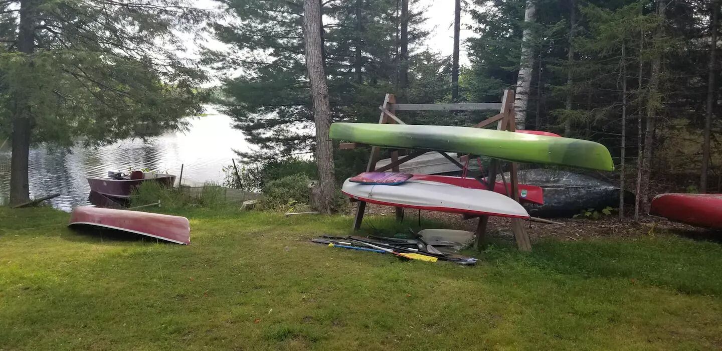 A grassy area with several canoes and kayaks, some stacked on a wooden structure, and a small boat docked by the water.