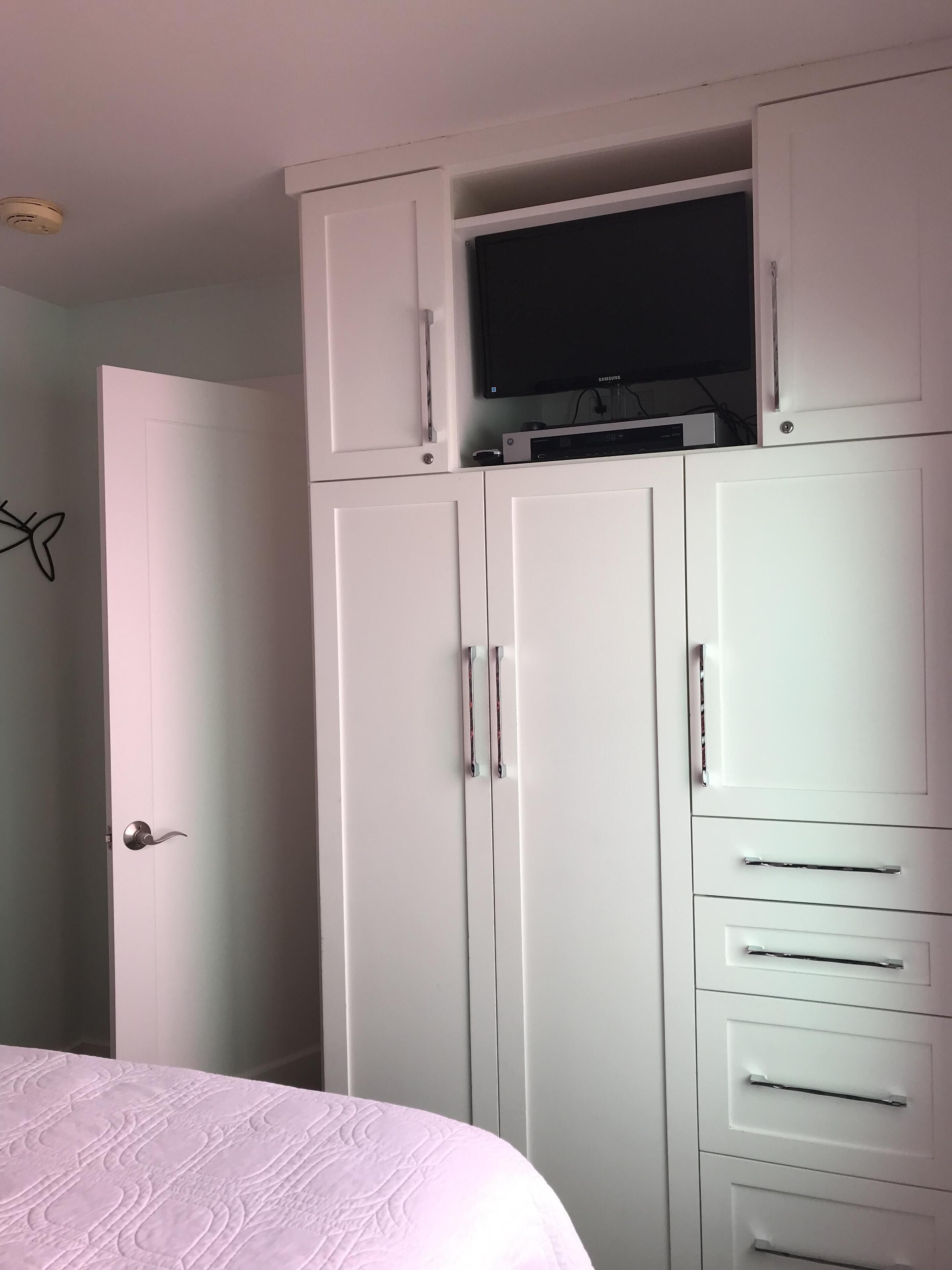 A bedroom with a bed, white wardrobe with drawers, a television on top, and a door.