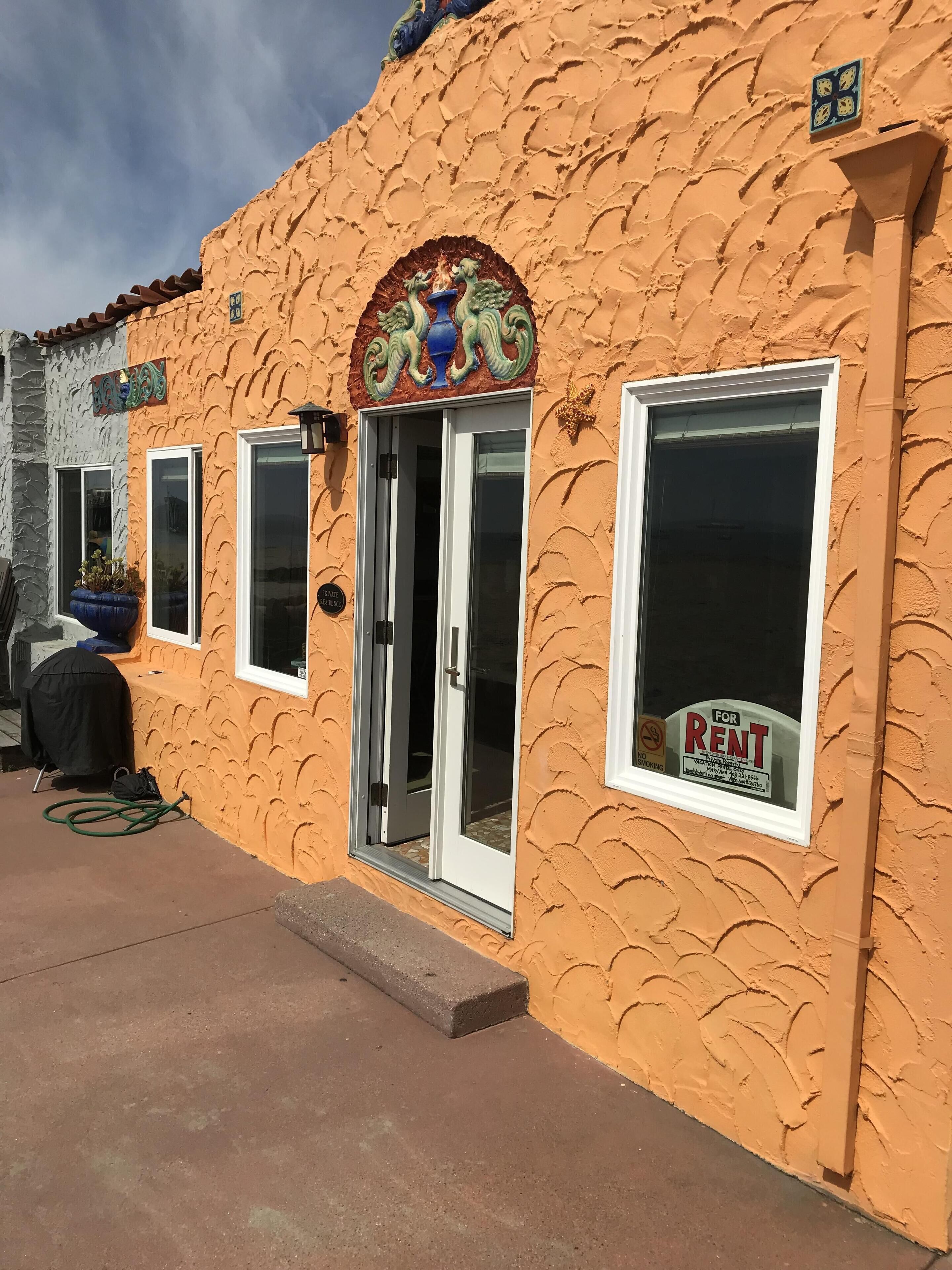 Un edificio con fachada naranja texturizada, una entrada decorativa con motivos de pavo real y un cartel de 'For Rent' en la ventana.