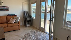 Un salon avec un canapé marron, un fauteuil, un sol carrelé et une vue sur une étendue d’eau, visible à travers une porte-fenêtre coulissante.
