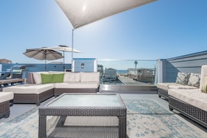 Eine Dachterrasse mit Korbmöbeln, einem Glastisch und Blick auf Strand und Meer.