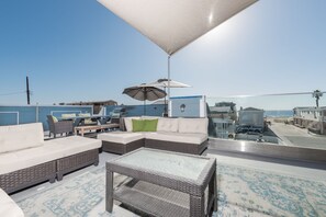 A rooftop patio with wicker furniture, a glass table, and a view of the ocean and buildings.