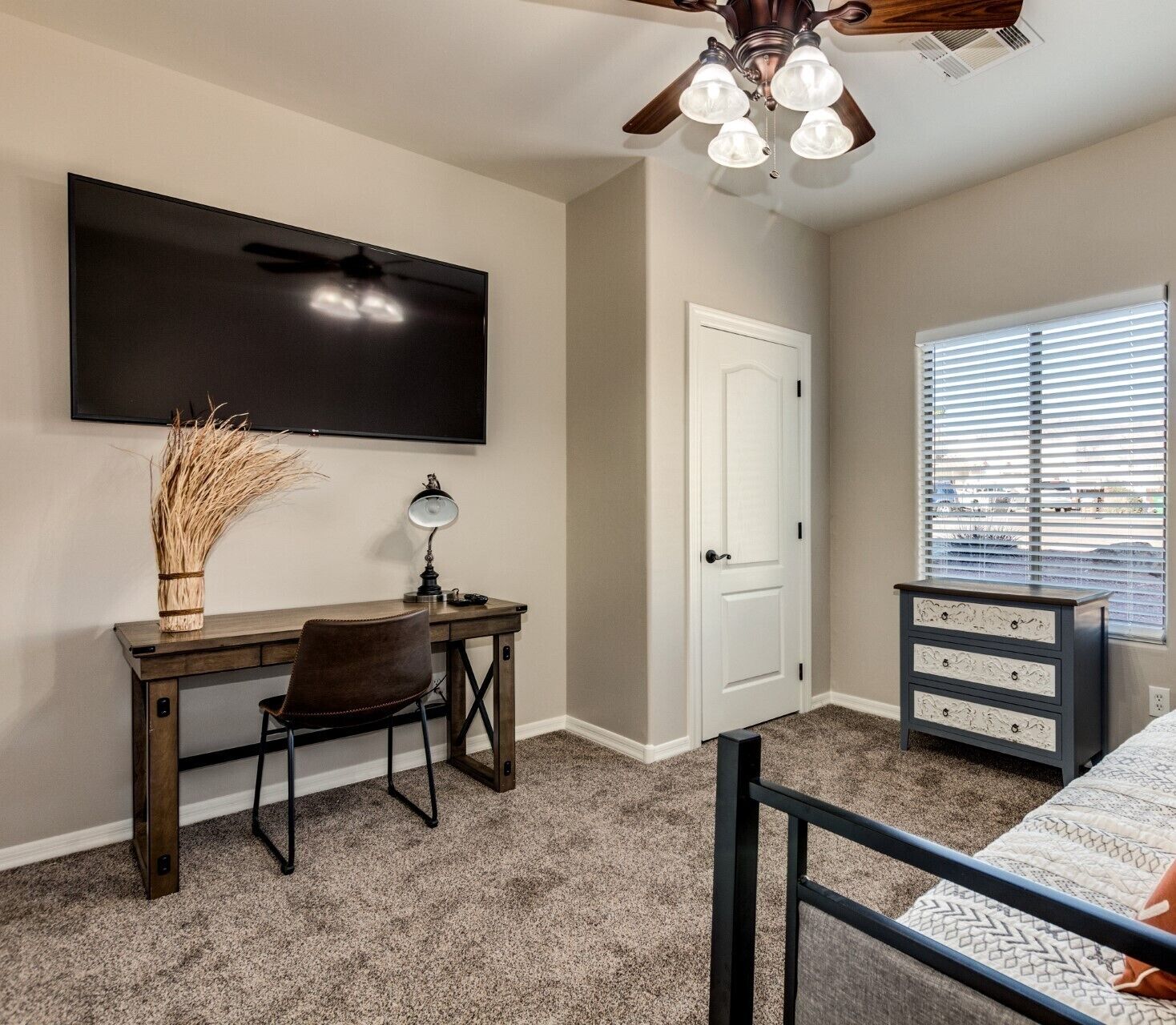 A bedroom with a bed, a desk with a chair, a TV, a ceiling fan, and a window with blinds.