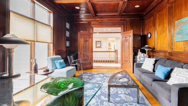 Un salon spacieux avec un plafond en bois foncé, un canapé gris agrémenté de coussins bleus, une table basse en verre et une grande fenêtre avec des stores.