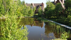 Ein friedlicher See mit einer Holzbrücke, üppigem Grün und traditionellen Gebäuden mit Schornsteinen.