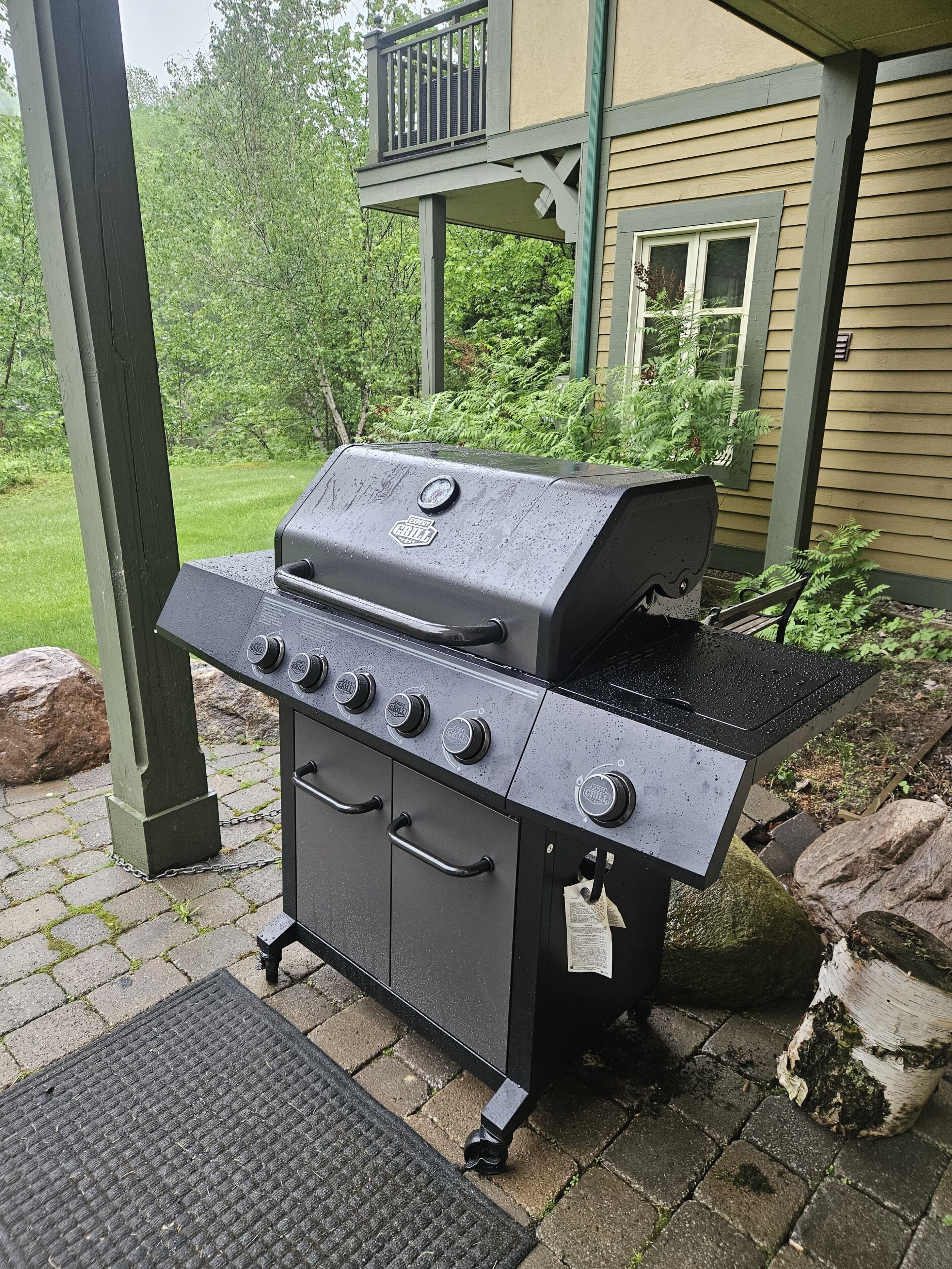 Una parrilla portátil con cuatro quemadores, un estante lateral y un panel de control, ubicada en un patio de ladrillo con cubierta de madera y vegetación de fondo.