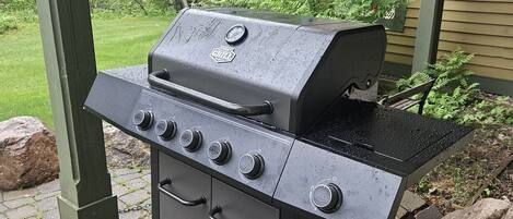 Una parrilla portátil con cuatro quemadores, un estante lateral y un panel de control, ubicada en un patio de ladrillo con cubierta de madera y vegetación de fondo.