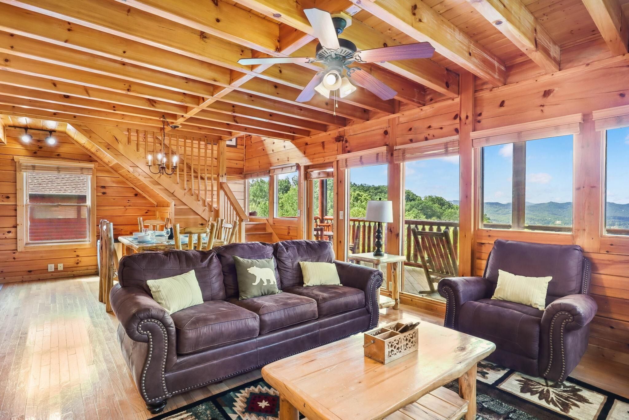 Interno di una baita in legno con un divano in pelle, poltrone, un tavolo da pranzo e una scala che conduce a un balcone con vista panoramica.