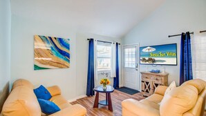 Un salón con dos sofás color beige, una mesa de centro y un cuadro en la pared. Hay una puerta que ofrece vistas al exterior, y un cartel que dice "Sierra Sun Villas".
