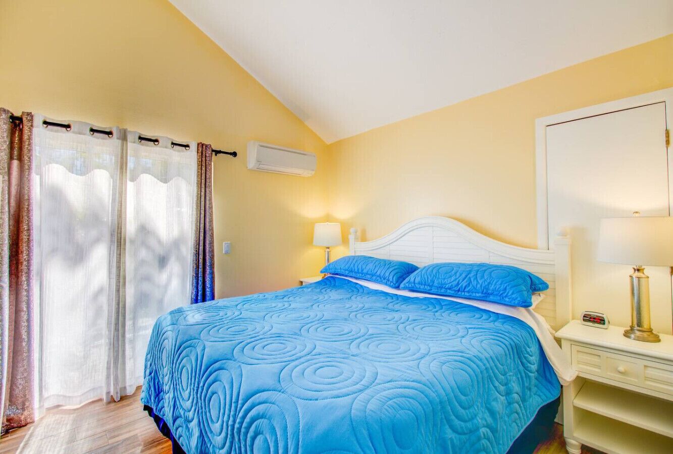 A bedroom with a bed, curtains, a nightstand, a lamp, and an air conditioning unit.