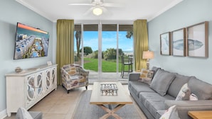 A spacious living room with a large TV, comfortable seating, a ceiling fan, and a view of the outdoors. - Waterscape B103 ~ 2BR Ground Floor ~ Gulf Views! (Fort Walton Beach)