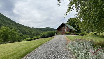 A log cabin with a gravel driveway leading to a wooden porch, surrounded by lush greenery and hills.