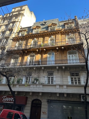 A multi-story building with ornate balconies, a closed shuttered storefront, and a visible tree in the foreground. - Luxury Restored Parisian Penthouse perfectly located to see all of Buenos Aires (Buenos Aires)