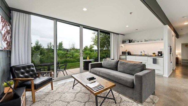 Un soggiorno moderno con un divano grigio, una poltrona in pelle nera e un tavolino da caffè in legno. Una porta scorrevole in vetro dà accesso a un balcone con vista sul verde.